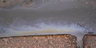 运行中的彩色雨水表面被汽油污染。石油污染。肮脏的彩虹水在城市街道的人行道旁流动