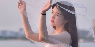 美丽的中国女孩在白色婚纱摆姿势，闭上眼睛和面纱在风的黄昏与模糊的河水背景，美丽，浪漫的概念，4k镜头，慢动作。