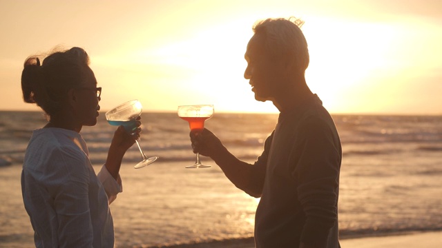 浪漫的亚洲老年夫妇看日落和享受一杯葡萄酒在美丽的热带海滩慢镜头。