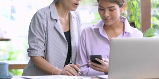 4K Medium拍摄的两位亚洲女商人在户外咖啡店使用智能手机和笔记本电脑讨论和分享金融业务的想法。户外放松的工作理念。