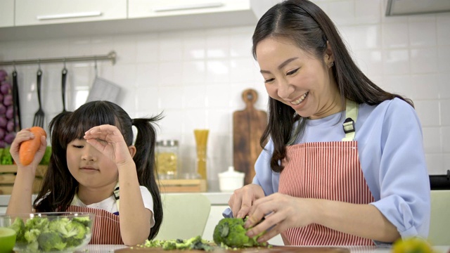 4k，年轻的亚洲母亲和女儿快乐和乐趣准备蔬菜和水果沙拉在一个现代化的白色厨房。女儿做饭时心情愉快。