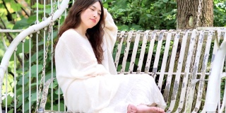 美丽的中国年轻女子穿着白色的裙子，光着脚坐在摇椅上摇摆睡觉，美丽的夏天，4k镜头，慢镜头。