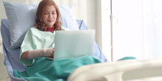 亚洲妇女生病的病人躺在医院的床上使用笔记本电脑放松时，她的病在医院恢复。