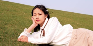 一名美丽的中国年轻女子躺在绿草如茵的草地上，手捧着头微笑着对着镜头，享受着春天的微风，4k镜头，慢镜头。