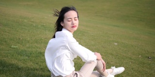 一个美丽的中国年轻女子坐在绿色的草地上，闭着眼睛，享受微风吹拂她的长发，4k视频，慢镜头。