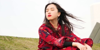 一个美丽的中国年轻女子坐在草地上，闭着眼睛，享受微风吹拂她的黑色长发，4k视频，慢镜头。