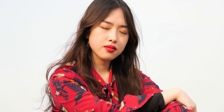 一个美丽的中国年轻女子坐在草地上，闭着眼睛，享受微风吹拂她的黑色长发，4k视频，慢镜头。