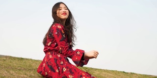 一名身穿红色连衣裙的美丽中国年轻女子坐在草地上，对着镜头微笑，享受微风吹拂她的黑色长发，4k视频，慢镜头。