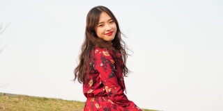 一名身穿红色连衣裙的美丽中国年轻女子坐在草地上，对着镜头微笑，享受微风吹拂她的黑色长发，4k视频，慢镜头。