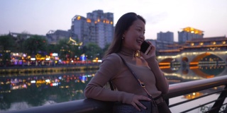 美丽的亚洲年轻女子戴着墨镜走在成都城市街道九烟桥的河边。美丽的长发女孩讲手机行走旅行在晚上中国滨江城市