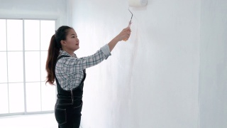 侧面的亚洲妇女与油漆滚筒油漆墙在她的新房子。视频素材模板下载