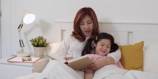 亚洲祖母在家给孙女读童话故事。年长的中国，奶奶高兴地放松与年轻女孩谁睡在卧室躺在床上听故事晚上在家里。缓慢的运动。