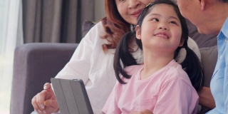 亚洲的祖父母和孙女在家里使用平板电脑。大四的中国，爷爷奶奶开心的花家人放松与年轻的女孩查看社交媒体，躺在客厅的沙发上。缓慢的运动。