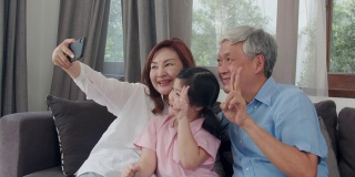 亚洲祖父母在家与孙女自拍。中国老年人，爷爷和奶奶愉快地度过家庭时间放松用手机和年轻的女孩孩子躺在客厅的沙发上。慢动作