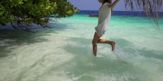 马尔代夫，一名身穿白色连衣裙的美丽年轻女子在碧绿的海水中奔跑。奇异的热带植物。缓慢的运动。