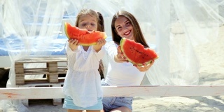 快乐快乐的小女孩穿着白色连衣裙和妈妈一起在海滩上吃西瓜。