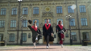 三名学生穿着黑色和红色的斗篷在大学大楼前庆祝他们的毕业。视频素材模板下载