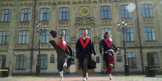 三名学生穿着黑色和红色的斗篷在大学大楼前庆祝他们的毕业。