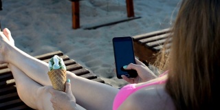 女孩穿着泳衣，坐在沙滩上的躺椅上用智能手机