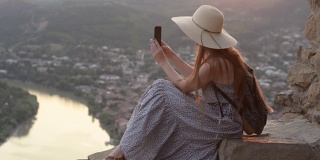 戴着帽子背着背包的女孩坐在山上。拍摄视频到智能手机，城市下面
