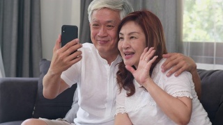 亚洲老年夫妇在家视频通话。中国老人躺在客厅的沙发上，用手机视频通话与家里的孙辈聊天。缓慢的运动。视频素材模板下载