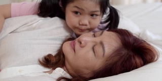 亚洲祖母在家睡觉。高三的中国，奶奶开心的放松与小孙女女孩亲吻脸颊为晚上醒来躺在卧室在家里的床上的概念。缓慢的运动。