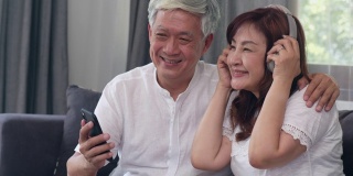亚洲老年夫妇在家里放松。亚洲老人中国祖父母，夫妻幸福戴着耳机用手机听音乐，躺在家里客厅的沙发上。缓慢的运动。