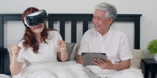 亚洲老年夫妇在家里使用平板电脑。亚洲资深的中国爷爷奶奶，在早上躺在家里卧室的床上一起玩虚拟现实游戏的概念。