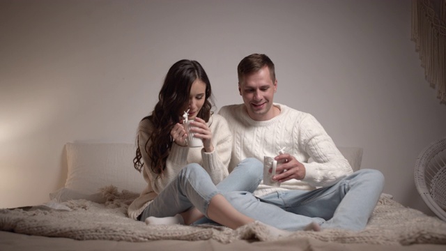 一对年轻夫妇坐在卧室的床上聊天。他们手里拿着一杯热饮，营造圣诞浪漫氛围。