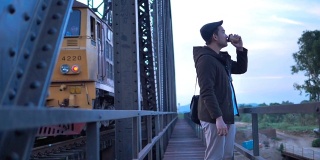 穿着大衣的亚洲游客在黑色铁桥铁路背景下喝着外卖咖啡杯。