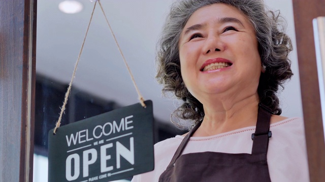 快乐的老年妇女拿着开放的牌子和欢迎邀请在咖啡店。创业、视觉、创新机会,小企业,老化,智慧,独立、协作、领导,退休,服务,Seniorpreneurs概念。