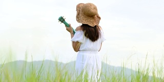 亚洲快乐的女人在白色的衣服弹奏尤克里里吉他在绿色的草地与山的背景在夏天。音乐家美女放松与乐器。乡村的生活方式