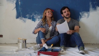 一对幸福的夫妇坐在地板上看着纸上的设计在家庭装修期间视频素材模板下载