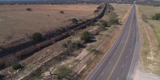 美国德克萨斯州爱德华兹高原上的一列货运火车正沿着一条高速公路行驶在稀树草原上。无人机用摄像机向前移动视频，跟踪列车。