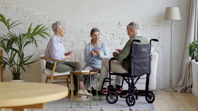 一组三个老人，两个坐在轮椅上的老年妇女和残疾老人，在养老院的公共休息室打牌，宽镜头