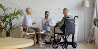 一组三个老人，两个坐在轮椅上的老年妇女和残疾老人，在养老院的公共休息室打牌，宽镜头