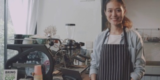 年轻美丽的亚洲女咖啡师穿着围裙，拿着热咖啡杯，微笑着在咖啡店的吧台招待顾客。咖啡店和小型咖啡店的概念。