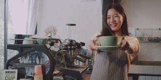 年轻美丽的亚洲女咖啡师穿着围裙，拿着热咖啡杯，微笑着在咖啡店的吧台招待顾客。咖啡店和小型咖啡店的概念。