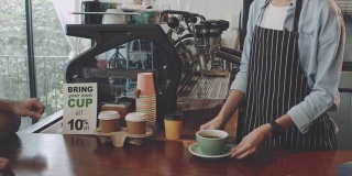 年轻美丽的亚洲女咖啡师穿着围裙拿着热咖啡杯在咖啡店的吧台服务给顾客。咖啡店和小型咖啡店的概念。