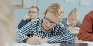 小学课堂:一个聪明的白人男孩戴着眼镜在练习本上写字。学习科学和创造性思维的多元聪明儿童群体
