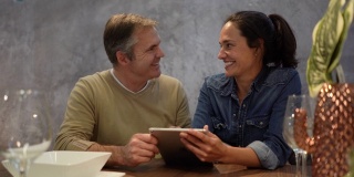 一对幸福的夫妇坐在家里的餐桌上一边聊天一边用平板电脑在网上购物