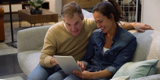 一对幸福的夫妇在家具店看着平板电脑上的产品，坐在沙发上又说又笑