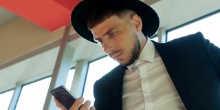 一名穿着黑色西装、戴着帽子的时髦白人男子手持手机，在机场看信息，特写，慢镜头