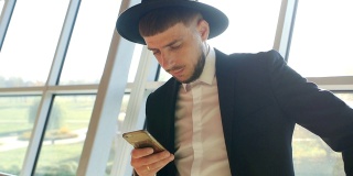 时尚的白人男子穿着黑色西装，戴着帽子，手里拿着手机，在机场看信息，特写，慢镜头