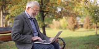 高级商人在外面使用笔记本电脑。一位身穿灰色夹克的老人在公园里使用笔记本电脑