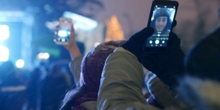 圣诞音乐会现场的人群在舞台附近用智能手机拍摄视频和自拍。年轻人在城市广场上举行新年晚会音乐剧表演。下雪的冬夜