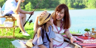 4K快乐的年轻亚洲家庭享受露营在湖。两位父母和可爱的小女孩一家人坐在帐篷前的露营椅上钓鱼和玩得一起在假期。