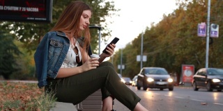年轻漂亮的女人坐在人行道上喝着咖啡，拿着手机找出租车