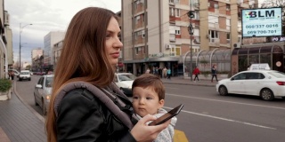 漂亮的年轻女子穿着黑色皮夹克和她的儿子在婴儿推车看她的智能手机和使用应用程序订出租车