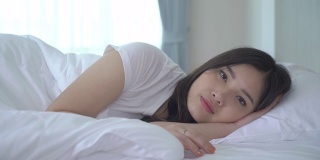 美丽的亚洲女人肖像与迷人的微笑享受新鲜柔软的床上用品亚麻布床垫在白色的卧室现代公寓。可爱的亚洲女孩睡觉休息，晚安睡眠的概念。
